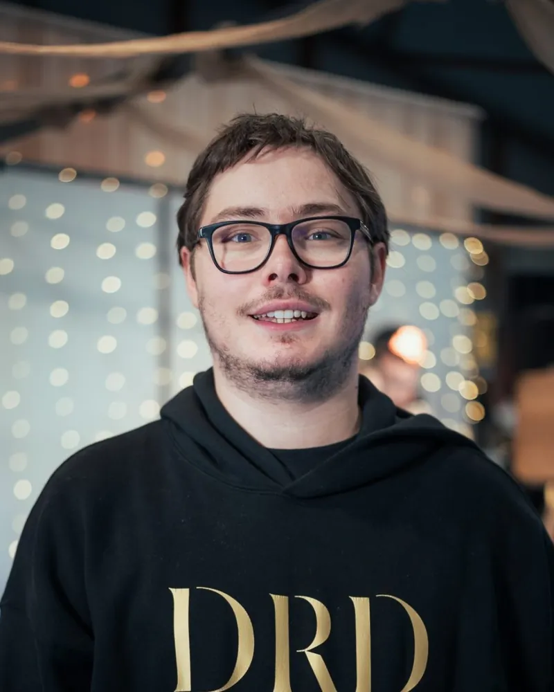 Adam souriant en sweat DRD Events devant un rideau lumineux